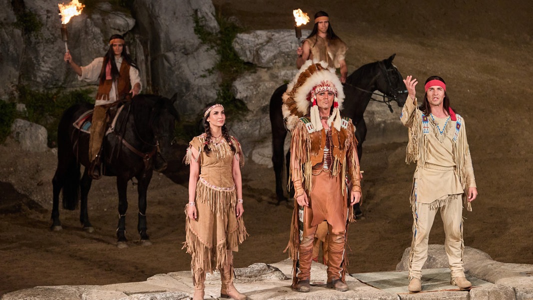 Im Vordergrund stehen drei Personen, verkleidet als Indigene. Dahinter sind zwei Reiter auf Pferden, auch als Indigene verkleidet, und halten brennende Fackeln hoch.