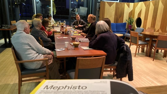 Menschen sitzen an einem langen Tisch und unterhalten sich. Ein Schriftstück im Vordergrund trägt die Überschrift "Mephisto". © NDR Foto: Janek Wiechers