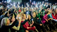 Viele Menschen blicken in einem großen Saal nach vorne und halten leuchtende Handys in die Höhe. © picture alliance/dpa | Christoph Soeder 