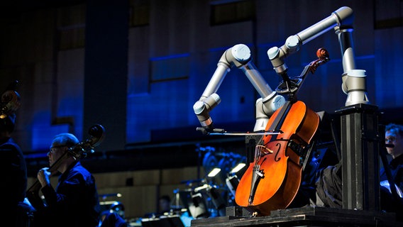 Ein Roboter, der Cello spielt, steht in einem Orchester auf einer Bühne. © Emma Larsson 