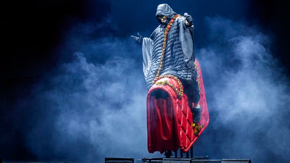 Auf einer Bühne sitzt ein Mann in einer langen Winterjacke auf einer überdimensionierten, roten Damenhandtasche. © picture alliance / Geisler-Fotopress | Michael Kremer/Geisler-Fotopress 