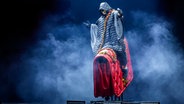 Auf einer Bühne sitzt ein Mann in einer langen Winterjacke auf einer überdimensionierten, roten Damenhandtasche. © picture alliance / Geisler-Fotopress | Michael Kremer/Geisler-Fotopress 