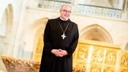 Ein Mann in einem schwarzen Gewand mit einem Kreuz um den Hals steht mit gefalteten Händen in einer Kirche und lächelt. © picture alliance/dpa | Hauke-Christian Dittrich 