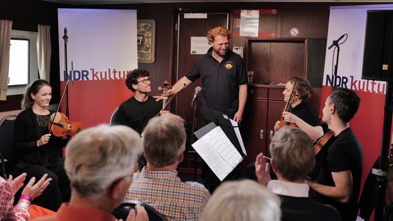 Julian Prégardien steht, umgeben von einem Streichquartett, in einer Schiffsmesse, in der Publikum sitzt. © NDR Foto: Cornelius Kob