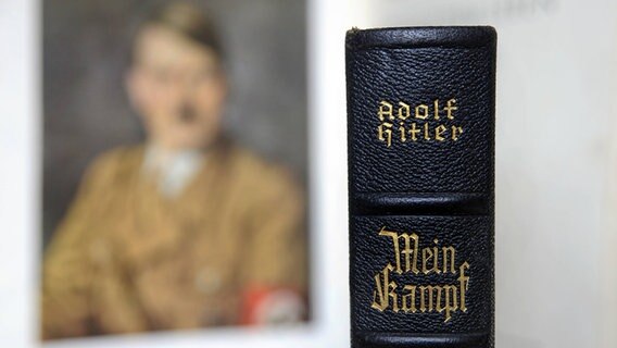 Auf einem Buchrücken steht: Adolf Hitler - Mein Kampf. Im Hintergrund hängt ein unscharfes Porträt Hitlers. © imago/sepp spiegl 