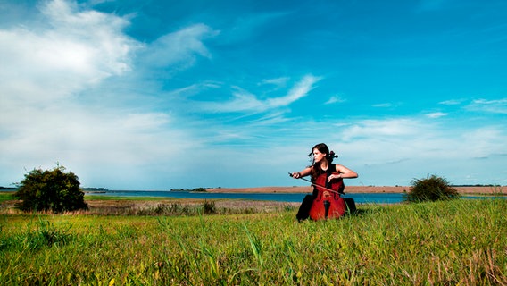 Eine Cellistin sitzt auf einer grünen Wiese vor blauem Himmel (Imagebild der Festspiele MV). © Felix Broede/Festspiele MV Foto: Felix Broede