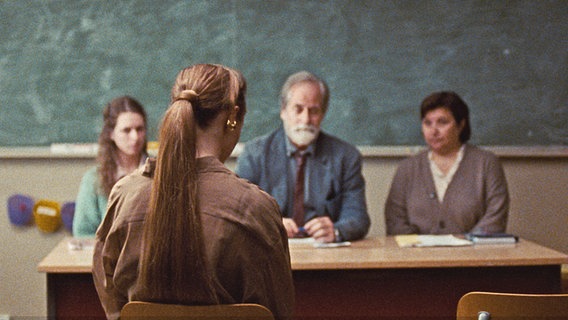 Eine Frau langem Pferdeschwanz sitzt in einem Klassenzimmer vor einem Pult, dahinter drei Personen - Szene aus "Armand" ©  Pandora Film / Eye Eye Pictures 