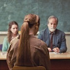 Eine Frau langem Pferdeschwanz sitzt in einem Klassenzimmer vor einem Pult, dahinter drei Personen - Szene aus "Armand" ©  Pandora Film / Eye Eye Pictures 