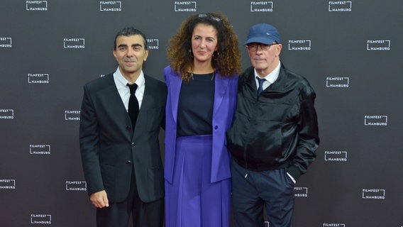 Zwei Männer in dunklen Anzügen flankieren eine Frau mit langen Haaren und einem lilafarbenen Anzug auf dem roten Teppich beim Filmfest Hamburg © NDR Foto: Patricia Batlle