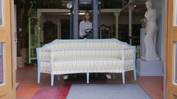 Eine Frau fährt ein antikes Sofa in einer Halle mit einem Gabelstapler  - Szene aus dem Dokumentarfilm "Die Ausstattung der Welt" © Bramkamp Weirich GbR 