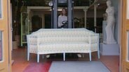 Eine Frau fährt ein antikes Sofa in einer Halle mit einem Gabelstapler  - Szene aus dem Dokumentarfilm "Die Ausstattung der Welt" © Bramkamp Weirich GbR 