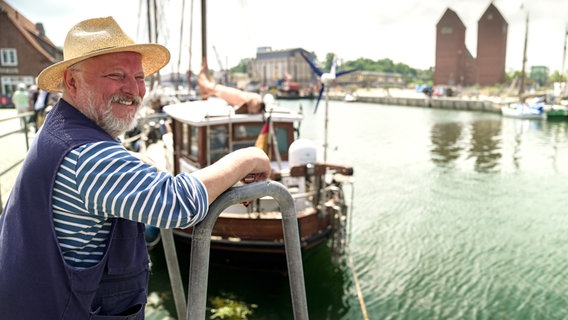 Ein Mann mittleren Alters mit Strohhut und weißblau gestreiftem Oberteil mit langen Armen steht lächelnd am Bord eines Schiffes, dahinter ist Wasser zu erkennen © WDR/Dokfilm/Jan Urbanski 