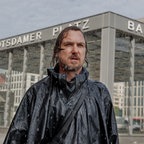 Ein Mann in grün gekleidet mit nassen Haaren steht im Regen an einer Berliner Kreuzung - Szene aus dme Film "Das Licht" © Frédéric Batier / X Verleih AG Foto: Frédéric Batier