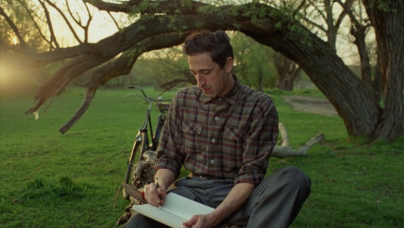 Ein Mann im Holzfällerhemd sitzt unter einem Baum und zeichnet - Adrien Brody im Film "Der Brutalist" von Brady Corbet © Universal Pictures 