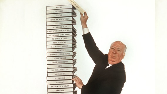 Alfred Hitchcock blickt in die Kamera und stapelt Drehbücher übereinander (Archivbild) © DB Bert Reisfeld/dpa +++ dpa-Bildfunk +++ Foto: Bert Reisfeld