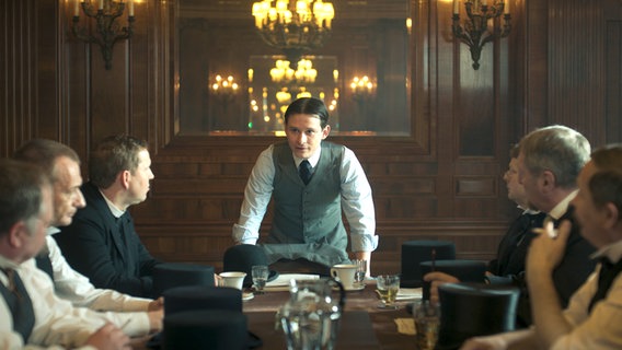 Franz Kafka (Joel Basman) spricht zu einer Gruppe von Männern, die an einem Tisch sitzen, in der ARD-Serie "Kafka". © NDR/Superfilm 