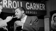 Szene aus dem Film "Misty - The Erroll Garner Story": Ein Mann zündet einem anderen eine Zigarette an. Im Hintergrund ist auf einer Werbetafel der Schriftzug "Erroll Garner" zu sehen. © 2024 FilmsGrandHuit 