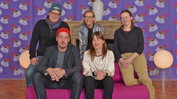 Ein Mann und eine Frau sitzen auf einem roten Sofa, dahinter hockt eine Frau seitlich und zwei weitere Personen stehen hinter dem Sofa und schauen frontal in die Kamera © Jasper Ehrich / Florian Arp Foto: Jasper Ehrich / Florian Arp