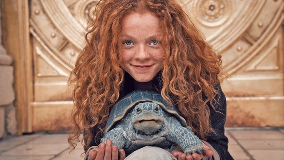 Ein Mädchen (Alexa Goodall) mit roten langen Locken und blauen Augen lächelt und hält eine Schildkröte - Szene aus "Momo" ein Kinofilm für 2025 © Constantin Film Verleih 