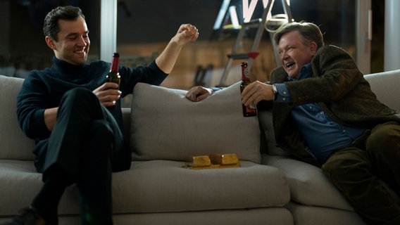 Der junge Anwalt Sven Lebert (Nils Strunk) und sein Chef Dr. Bernd Hausner (Justus von Dohnányi) sitzen auf einem Sofa und prosten sich freuend mit einer Flasche Bier zu. © X Filme Creative Pool GmbH / True Content Entertainment ApS / EPO-Film / ZDF / DR / Petro Domenigg 