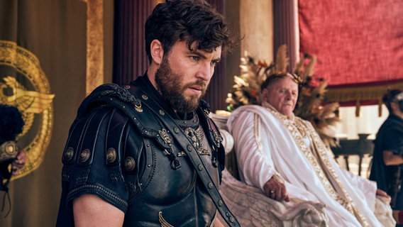 Tom Hughes als Titus im Vordergrund und im Hintergrund Anthony Hopkins als Kaiser Vespasian auf einem Thron. © Peacock Foto: Reiner Bajo