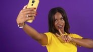 Studio-Porträt mit lila Hintergrund eines afrikanischen städtischen Frau gestikulieren Erfolg bei der Aufnahme eines Selfie mit Handy © picture alliance / imageBROKER | Unai Huizi 