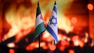 Die israelische Flagge und die palästinensische Flagge stehen auf einem Tisch, im Hintergrund ist Feuer zu sehen. © imago 