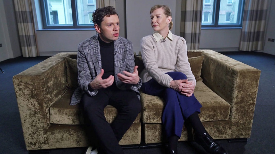 Christian Friedel und Sandra Hüller sitzen auf einem grau-braunen Sofa.