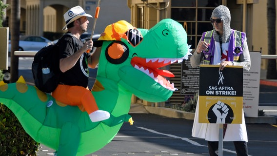 Zwei verkleidete Schauspieler in Dinosaurier- und Ritterkostüm stehen mit Protestplakaten auf der Straße. © picture alliance / ZUMAPRESS.com | Gene Blevins 