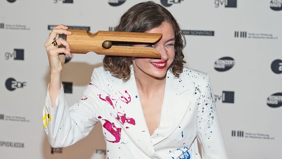 Die Cellistin Anastasia Kobekina mit kurzen lockigen Haaren und weißem Blazer mit buntem Flecken hält den Echo Klassik Award in einer Hand und schaut mit einem Auge durch © Picture alliance / ABBfoto 