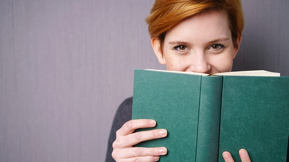 Junge Frau lächelt hinter einem Buch hervor. © fotolia.com Foto: contrastwerkstatt