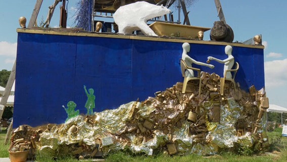 Vor einer blauen Wand türmt sich goldfarbener Müll © Screenshot NDR 