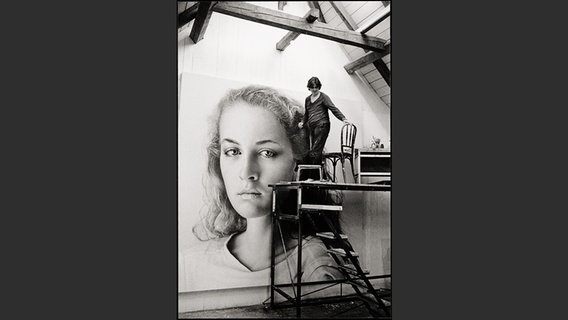 Ein Schwarz-Weiß-Foto in dem ein Maler an einem großformatigem Bild einer jungen Frau mal  - Franz Gertsch auf einer Leiter malt "Johanna" © Franz Gertsch AG. Foto: Balthasar Burkhard