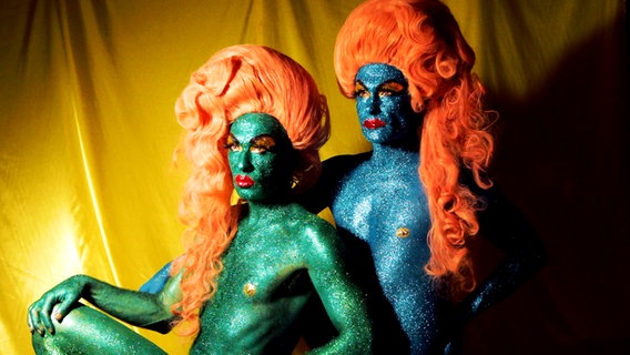Zwei vollkommen mit blauer, beziehungsweise grüner Glitzerfarbe bemalte Personen mit langhaarigen Perücken posen vor der Kamera. © The Huxleys 2021 Foto: The Huxleys