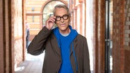 Ein Mann mit blauem Pulli, dunklem Sakko und dunkler Brille lächelt mit der Hand an die Brille gehoben - Wolfgang Joop © Christophe Gateau/dpa +++ dpa-Bildfunk +++ Foto: Christophe Gateau