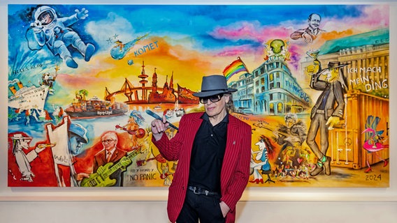 Udo Lindenberg steht mit einem Pinsel in der Hand vor seinem bislang größten Gemälde, das verschiedene Motive aus seiner Musik und der Schifffahrt zeigt. © Tine Acke/Hapag-Lloyd Presseteam /dpa 