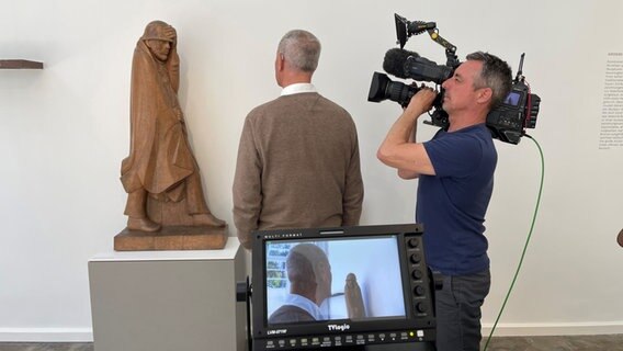 Ein Mann betrachtet eine Skulptur aus Holz, daneben steht ein Kameramann, der ihn filmt. © NDR Foto: Thomas Naedler