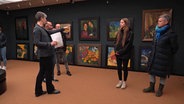 Besucher stehen in einem Ausstellungsraum des Museums vor einer Wand an der bunte Bilder hängen. © NDR Screenshot Foto: NDR Screenshot