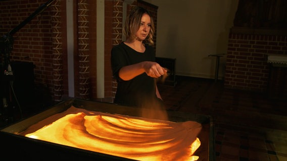 Irina Titova streut Sand auf eine von hinten beleuchtete Glasscheibe. © NDR 