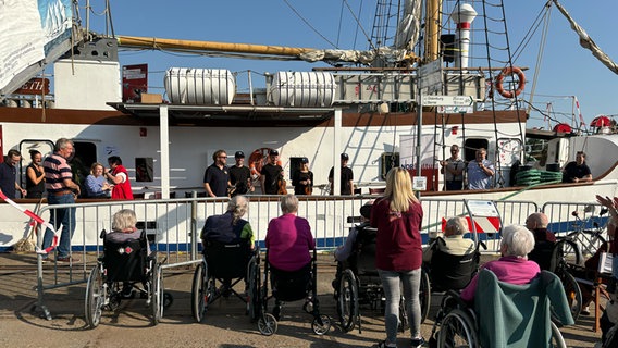 Eine Reihe aus Senior*innen in Rollstühlen sitzen auf dem Kai vor dem Schiff. Im Hintergrund stehen die Musizierenden an der Reling und sprechen mit den Gästen. © NDR Foto: Tom Holste