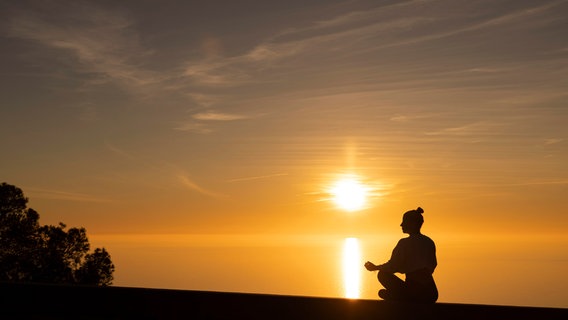 Eine Frau meditiert im Sonnenuntergang am Meer. © picture alliance / Westend61 | DREAMSTO 