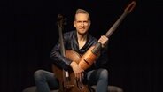 Der Cellist Johannes Moser sitzt vor zwei Instrumenten, einem akkustischem und einem elektrischem Cello. Dabei trägt er eine schwarze Lederjacke und hat blonde, kurze Haare. Er sitzt vor einem schwarzen Hintergrund. © Johannes Moser 