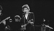 Der Sänger Bob Dylan steht auf einer Bühne mit einer Gitarre in der Hand, er singt in ein Mikrofon. © picture alliance / AP Images | Jeff Robbins Foto: picture alliance / AP Images | Jeff Robbins