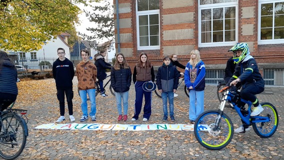 Sechs Schülerinnen und Schüler stehen auf dem Schulhof und werden von Kindern auf Fahrrädern umkreist. Vor ihnen liegt Papier mit der Aufschrift "Augustinus" © NDR 
