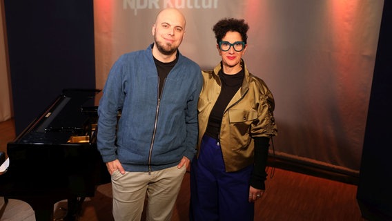 Ein Mann mit Glatze und einem Bart steht lächelnd neben einer Frau mit dunklen, kurzen Locken und einer großen, blauen Brille. Sie beide stehen vor einem schwarzen Flügel. © Ekaterina Shurygina / NDR Foto: Ekaterina Shurygina / NDR