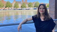 Eine Frau mit schwarzem Oberteil lehnt an einem Geländer am Fluss. Im Hintergrund ist die Altstadt von Lübeck zu sehen. © NDR Foto: Linda Ebener