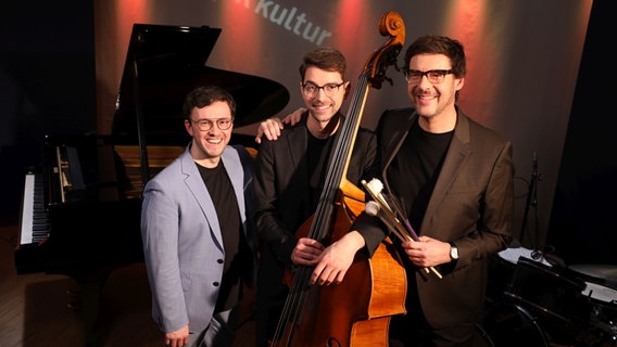 Drei Männer stehen lachend nebeneinander vor einem schwarzen Flügel. Der Mann in der Mitte hält einen Bass in der Hand und der Mann rechts Schlegel. © Ekaterina Shurygina / NDR Foto: Ekaterina Shurygina / NDR