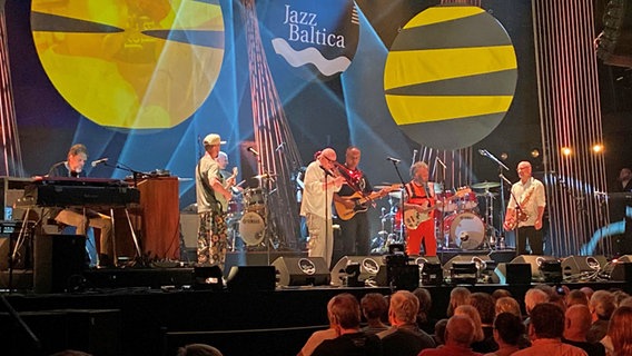 Auf einer Bühne mit der Aufschrift "JazzBaltica" stehen acht Musiker mit verschiedenen Instrumenten. © NDR / Linda Ebener Foto: Linda Ebener