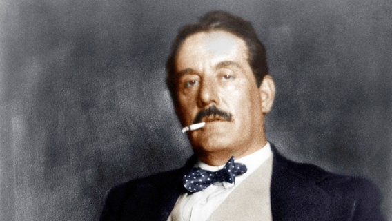 Giacomo Puccini in einem Anzug mit blauer Fliege und weißen Punkten. In seinem Mund, von seinen Lippen gehalten, hat er eine Zigarette und befindet sich vor einem dunklen Hintergrund. © picture alliance 