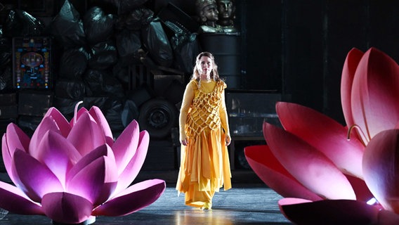 Lea-ann Dunbar in der Hauptrolle der 'Sita' steht in einem gelben Kleid auf der Bühne. Links und rechts von ihr befindet sich jeweils eine große pinkfarbende Blüte. © Martin Kaufhold Foto: Staatstheater Saarbrücken